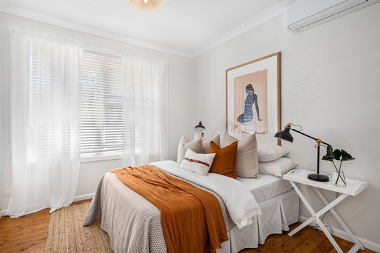The neutral linen bed is accented with orange throw pillows and blanket, paired with warm wood tones and sleek black and gold light fixtures.