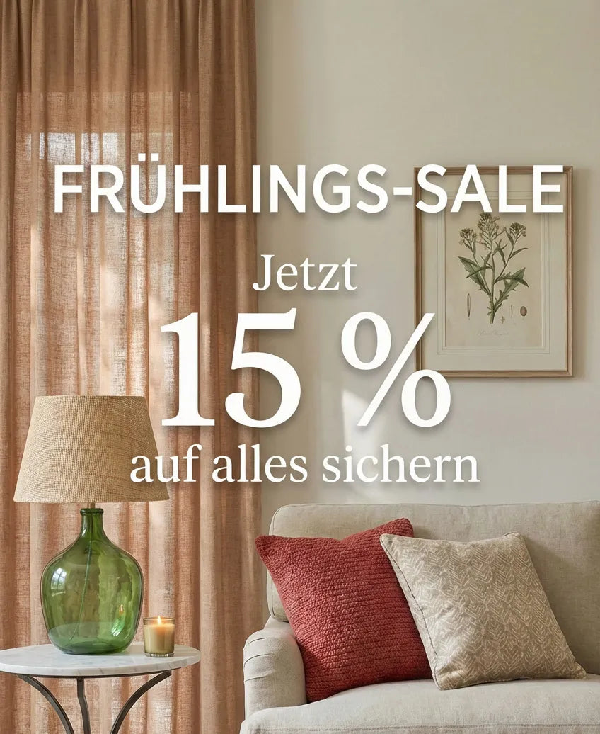 Ein Wohnzimmer im Innenbereich mit einem Sofa, einem kleinen Beistelltisch, Vorhängen und gerahmter Kunst an der Wand. Der Beistelltisch hat eine grüne Glaslampe mit einem gewebten Schirm und einer kleinen brennenden Kerze daneben. Der Text auf dem Bild lautet: FRÜHLINGS-SALE Jetzt 15 % auf alles sichern